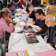 Várias pessoas estão sentadas frente a frente em mesas compridas cobertas por toalhas brancas. Em cada mesa há notebooks, documentos e canetas. Os atendentes, posicionados de um lado das mesas, digitam ou analisam papéis enquanto conversam com as pessoas atendidas. A imagem mostra um ambiente amplo, semelhante a um ginásio, com piso verde e algumas faixas de isolamento e cones laranja ao fundo. As pessoas utilizam roupas casuais e o local está movimentado, com várias fileiras de atendimento.
