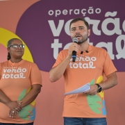 Uma foto em close-up, capturada em um evento interno, mostra um grupo de quatro pessoas paradas em frente a uma parede de fundo colorida que promove a "operação verão total".O foco principal está em um homem de pele clara no centro-direita, que está falando em um microfone que segura com a mão direita. Ele veste uma camiseta laranja promocional com o logotipo "operação verão total" em letras brancas. Ele segura um pedaço de papel dobrado na mão esquerda e usa um relógio de pulso preto.Ao lado dele, no centro-esquerda, está uma mulher de pele escura com cabelo curto e óculos. Ela também veste a mesma camiseta laranja promocional e tem os braços cruzados na frente.Outros dois homens aparecem parcialmente:À esquerda, um homem de pele clara com uma camiseta escura.À extrema direita, um homem de pele clara em uma camisa cinza-escura.O fundo é um painel de parede com as palavras "operação verão total" escritas em branco em grandes faixas horizontais de cores laranja, roxo e verde-água. A faixa roxa, mais proeminente, contém a palavra "verão" e o til ($\tilde{}$) alongado.