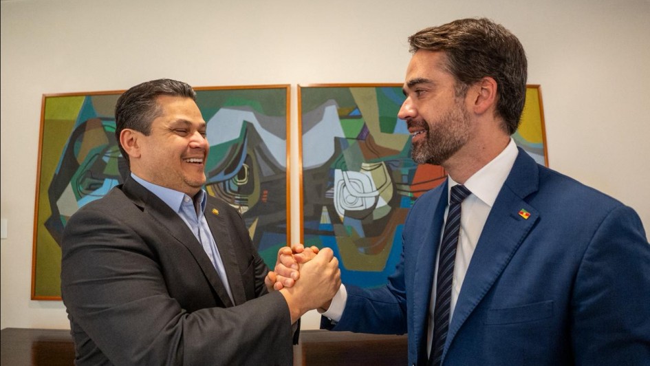 Foto em plano americano de Davi e Leite de lado. Eles estão um de frente para o outro e se dão as mãos, sorrindo.