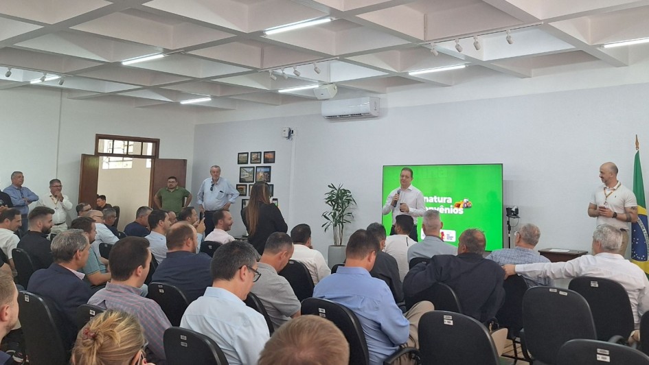 Foto mostra secretário da Agricultura, em pé em frente à um monitor que mostra uma tela verde. Ele está falando com microfone na mão, em uma sala com paredes brancas. Algumas pessoas estão em pé. A plateia está cheia, com muitos homens sentados de costa para a foto, olhando a fala do secretário.