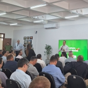 Foto mostra secretário da Agricultura, em pé em frente à um monitor que mostra uma tela verde. Ele está falando com microfone na mão, em uma sala com paredes brancas. Algumas pessoas estão em pé. A plateia está cheia, com muitos homens sentados de costa para a foto, olhando a fala do secretário.