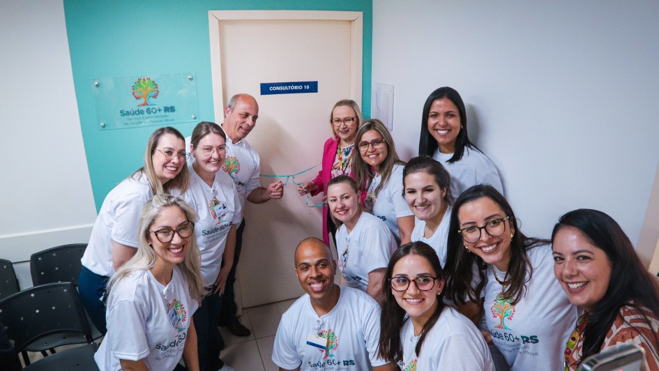 A imagem mostra um grupo de pessoas vestindo camisetas do programa Saúde 60+ RS em frente a uma porta de consultório, posando para a foto. Na parece, há uma placa do programa.