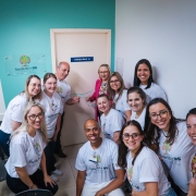 A imagem mostra um grupo de pessoas vestindo camisetas do programa Saúde 60+ RS em frente a uma porta de consultório, posando para a foto. Na parece, há uma placa do programa.