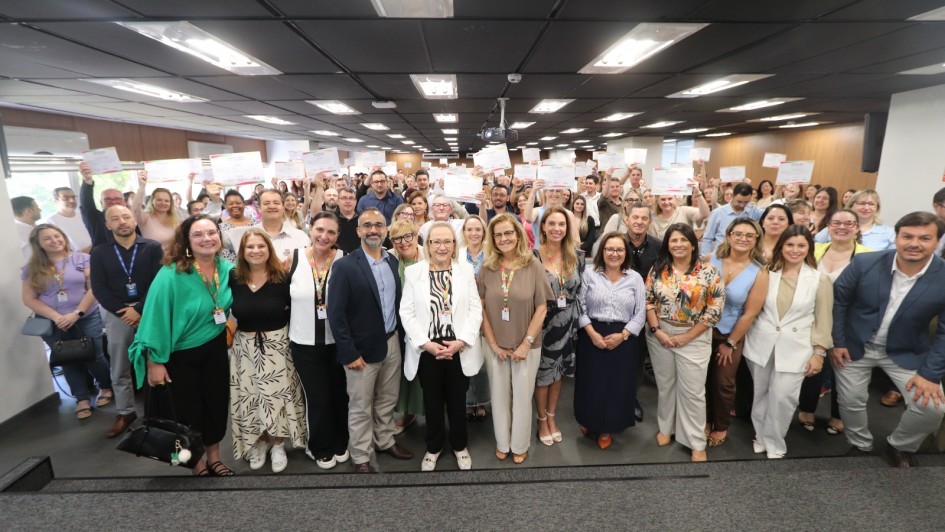 Descri&ccedil;&atilde;o da Imagem para Acessibilidade
Uma foto de plano aberto tirada em um audit&oacute;rio interno mostra um grande grupo de pessoas reunidas para uma cerim&ocirc;nia ou evento de encerramento, provavelmente relacionado ao "Qualifica Vigil&acirc;ncia RS 2025".

Pessoas:
Composi&ccedil;&atilde;o: O grupo &eacute; composto por uma grande quantidade de homens e mulheres, de diversas idades, alinhados em v&aacute;rias filas, ocupando a largura do ambiente. H&aacute; uma mistura de vestimentas casuais e sociais.

A&ccedil;&atilde;o Principal: Quase todas as pessoas no fundo e nas laterais est&atilde;o segurando certificados ou folhas de papel branco acima da cabe&ccedil;a com as duas m&atilde;os, em um gesto de celebra&ccedil;&atilde;o ou conclus&atilde;o de um curso.

Foco Central: Na primeira fila, no centro, h&aacute; um grupo de cerca de 10 a 12 pessoas que parecem ser l&iacute;deres ou organizadores. Entre elas, uma mulher de cabelos loiros e um casaco branco e preto se destaca no centro. Outras mulheres na frente vestem roupas coloridas e saias longas.

Cen&aacute;rio:
Teto: O teto &eacute; baixo e forrado com diversas lumin&aacute;rias retangulares de LED, que iluminam bem o ambiente.

Paredes: As paredes laterais s&atilde;o neutras, com janelas no lado esquerdo que deixam entrar alguma luz natural.

Ch&atilde;o: O ch&atilde;o &eacute; revestido com carpete cinza escuro.

Estrutura: O grupo est&aacute; em p&eacute; em uma pequena eleva&ccedil;&atilde;o ou estrado forrado, indicando que est&atilde;o em uma &aacute;rea de palco ou de apresenta&ccedil;&atilde;o.