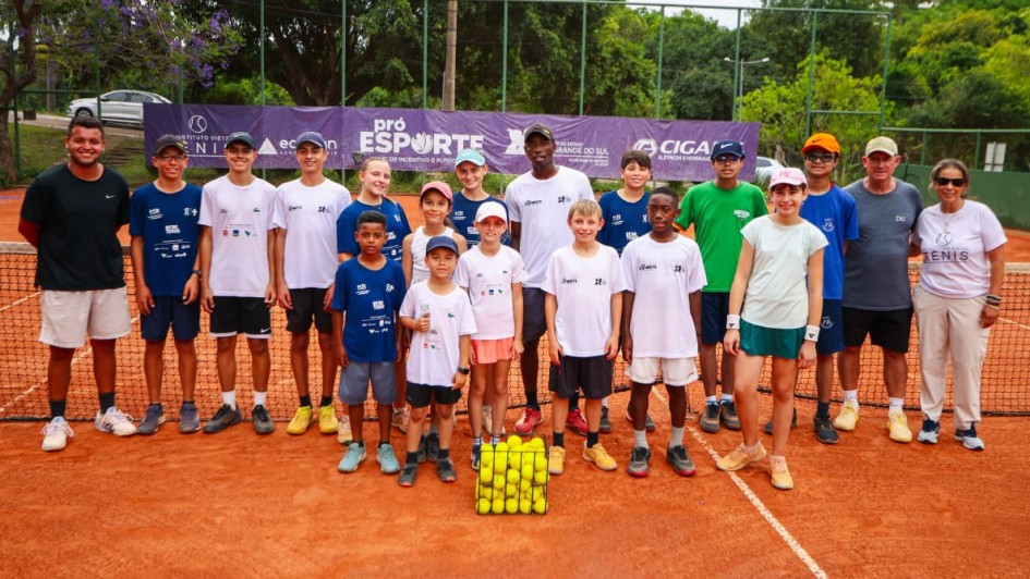 Um grupo de aproximadamente vinte pessoas, incluindo crianças, adolescentes e adultos, está reunido em uma quadra de saibro de tênis. Todos aparecem alinhados em duas fileiras diante da rede. A maior parte veste camisetas esportivas brancas ou azuis, e alguns usam bonés. No centro, à frente, há uma pilha organizada de bolas de tênis amarelas. Ao fundo, um banner roxo com texto referente ao programa “Pró-Esporte” está preso à grade de proteção da quadra. Árvores verdes e uma rua com um carro aparecem atrás da grade.