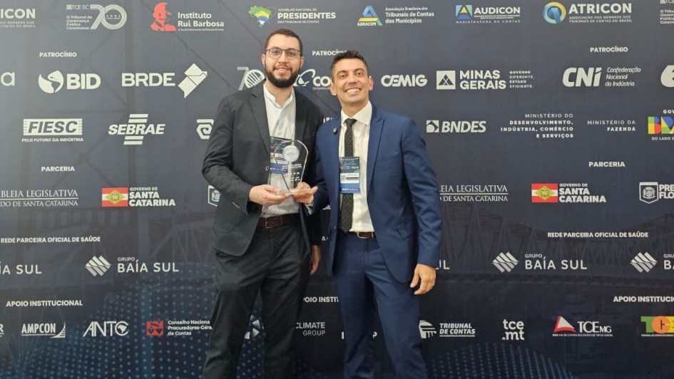 Foto mostra dois homens posando para foto, em frente a um painel de fundo preto com diversas logomarcas e o s&iacute;mbolo do evento. Um deles est&aacute; com um trof&eacute;u de acr&iacute;lico.
