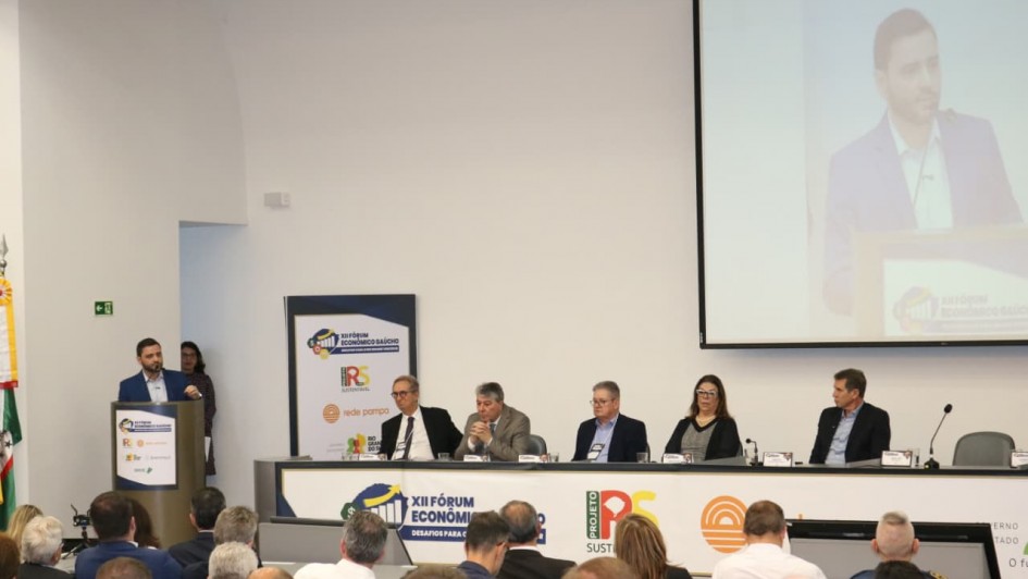 Uma foto colorida e em plano geral registra uma mesa de debate durante o "XII Fórum Econômico Gaúcho", conforme indicado pelos logotipos e painéis no ambiente.

No centro da imagem, um palestrante, um homem branco com barba e terno azul-marinho, está de pé em um púlpito à esquerda. Ele gesticula enquanto fala para uma plateia sentada na parte inferior da imagem. No fundo e à direita, um telão reproduz o close-up do palestrante.

À direita do palestrante, sentados à mesa, estão cinco debatedores (quatro homens e uma mulher) atentos ao discurso. No painel à frente da mesa, lê-se "XII FÓRUM ECONÔMICO GAÚCHO - DESAFIOS PARA O FUTURO".

A sala tem paredes brancas, e a plateia é composta por pessoas de costas, majoritariamente em trajes sociais.