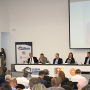 Uma foto colorida e em plano geral registra uma mesa de debate durante o "XII Fórum Econômico Gaúcho", conforme indicado pelos logotipos e painéis no ambiente.

No centro da imagem, um palestrante, um homem branco com barba e terno azul-marinho, está de pé em um púlpito à esquerda. Ele gesticula enquanto fala para uma plateia sentada na parte inferior da imagem. No fundo e à direita, um telão reproduz o close-up do palestrante.

À direita do palestrante, sentados à mesa, estão cinco debatedores (quatro homens e uma mulher) atentos ao discurso. No painel à frente da mesa, lê-se "XII FÓRUM ECONÔMICO GAÚCHO - DESAFIOS PARA O FUTURO".

A sala tem paredes brancas, e a plateia é composta por pessoas de costas, majoritariamente em trajes sociais.