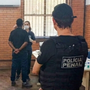 A foto mostra uma sala com paredes de tijolos aparentes e janelas amplas com persianas. No centro, uma pessoa em pé, de costas para a câmera, está com as mãos cruzadas atrás do corpo enquanto conversa com uma profissional de saúde que usa máscara e luvas, posicionada diante de uma mesa com materiais de coleta. À direita, um agente penal aparece em primeiro plano, também de costas, vestindo colete tático com a inscrição “POLÍCIA PENAL”. No canto da sala há uma cadeira azul e alguns itens sobre a mesa. O ambiente é iluminado pela luz natural que entra pelas janelas.