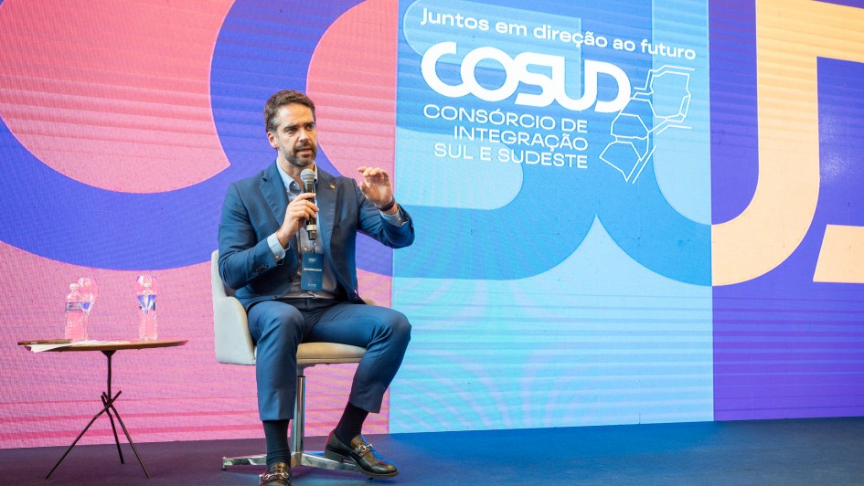 O governador Eduardo Leite está sentado em uma cadeira clara no palco, segurando um microfone na mão direita enquanto faz um gesto com a mão esquerda. Ele veste terno azul e sapatos escuros. À esquerda, há uma pequena mesa com duas garrafas de água. Ao fundo, um grande painel colorido exibe formas geométricas em tons de azul, rosa e amarelo, com o texto: “Juntos em direção ao futuro — COSUD — Consórcio de Integração Sul e Sudeste”, acompanhado de um desenho estilizado de mapas dos estados. O piso do palco é escuro e o cenário tem iluminação homogênea.