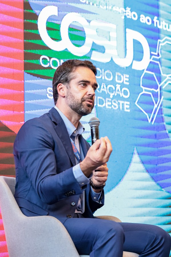 A foto mostra o governador Eduardo Leite em uma cadeira clara, segurando um microfone com a mão direita enquanto faz um gesto com a mão esquerda, com os dedos próximos. Ele veste terno azul e camisa social clara.Ao fundo, há um grande painel digital com design colorido em tons de azul, rosa e verde, contendo o texto “COSUD – Consórcio de Integração Sul e Sudeste”. 