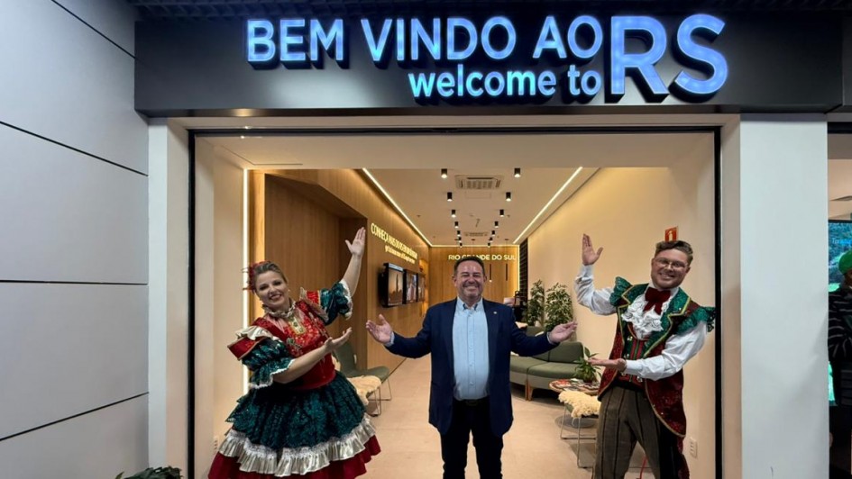 Uma fotografia em plano médio registra três pessoas posando na entrada de um espaço comercial ou informativo, provavelmente em um aeroporto, devido ao contexto do texto na placa superior.

Pessoas e Posições
Centro: Um homem branco, de meia-idade, com cabelos curtos e vestindo um blazer azul-claro sobre uma camisa de botão (sem gravata), está posicionado no centro da entrada. Ele tem uma expressão sorridente e os braços abertos em um gesto de boas-vindas.

Esquerda (Traje Típico): Uma mulher branca, de cabelos loiros parcialmente presos, veste um traje folclórico de cor predominante verde-escura e vermelho, com detalhes dourados e rendas. Ela está com os braços erguidos, com uma expressão animada, posando para a foto.

Direita (Traje Típico): Um homem branco, de óculos, bigode e cabelo curto, veste um traje folclórico masculino com colete em cores bordô e dourado, camisa branca com babados e calças xadrez/listradas em tons terrosos. Ele também está com os braços erguidos em uma pose de boas-vindas.

Fundo/Canto: No canto direito, parte de outra pessoa, vestindo um boné verde e uma jaqueta escura, é visível, possivelmente um turista.

Cenário e Decoração
Placa de Entrada: Acima do vão de entrada, uma placa luminosa em destaque diz: "BEM VINDO AO R S" e, abaixo, em inglês e letras menores, "welcome to". O "RS" está em azul e em negrito.

Interior do Espaço: O interior da sala é bem iluminado, com iluminação embutida no teto. Paredes e móveis (incluindo sofás e plantas no fundo) são em tons neutros e amadeirados, sugerindo um lounge ou centro de informações.

Piso: O piso da área externa é em porcelanato claro e liso.

Contexto: A placa de boas-vindas em português e inglês, a presença de pessoas com trajes típicos e a sugestão de aeroporto (com base no título da manchete) reforçam o tema de recepção a turistas no Rio Grande do Sul (RS).