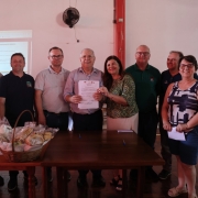Oito pessoas estão posicionadas atrás de uma mesa de madeira. No centro, um homem segura um documento impresso, com uma mulher ao lado apoiando o papel com uma das mãos. Os demais estão alinhados, olhando para a câmera. Sobre a mesa, há uma cesta com pães e biscoitos embalados, além de garrafas de água, copos, documentos e canetas. Ao fundo, uma projeção é exibida na parede branca, com textos parcialmente visíveis.