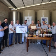 Uma fotografia em plano geral registra um grupo de treze pessoas, majoritariamente homens, posando em torno de uma mesa com garrafas de vinho, em um ambiente interno que se assemelha a uma vinícola ou agroindústria. O grupo celebra o recebimento de uma certificação ou premiação.

Pessoas e Posições
O grupo é composto por treze pessoas (onze homens e duas mulheres) vestindo, em sua maioria, trajes casuais.

Centro-Esquerda: Um pequeno grupo de homens está à esquerda, com um deles (o segundo da esquerda) segurando um certificado ou placa azul e branco que parece ser uma premiação. Outros homens próximos a ele estão batendo palmas.

Centro-Direita: Um homem mais velho, no centro, segura um certificado em papel pardo (cor de papelão) com as duas mãos, posando orgulhosamente. Uma mulher e um homem mais jovens estão atrás dele.

Extremidades: O grupo se estende horizontalmente, com a maioria das pessoas sorrindo e olhando para a câmera.

Cenário e Decoração
Ambiente Industrial: O local parece ser o interior de um armazém ou área de processamento. O chão é de concreto, as paredes são de tijolos à vista (cimento queimado/alvenaria bruta), e o teto é metálico com telhas de zinco.

Tanques: No fundo, visíveis à esquerda, há grandes tanques cilíndricos, provavelmente de aço inoxidável e fibra (ou concreto revestido) utilizados no processo de fermentação ou armazenamento de vinho.

Mesa de Exposição: No centro, há uma rústica mesa de madeira, sobre a qual estão dispostas diversas garrafas de vinho, frutas (como pêssegos e laranjas) e alguns petiscos ou alimentos.

Textos Visíveis (em Banners e Certificados)
Um banner suspenso no fundo, à esquerda, apresenta o texto em destaque: "Natureza do Bio" e o logotipo da EMATER-RS aparece em outro banner menor e também no certificado azul/branco, que diz "AQUI TEM EMATER/RS".

A foto transmite uma sensação de sucesso, parceria e celebração da produção agroindustrial.