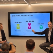 A imagem é uma fotografia de uma apresentação que mostra um painel digital (tela) com o título "CARTEIRA DE PROJETOS - PROJETOS TRABALHADOS PELA EQUIPE INVEST RS".

Elementos da Cena:
Tela de Apresentação (Plano Central):

R$ 10.9 BI (Seta Amarela): Indica "Em novos investimentos captados pela Invest RS".

R$ 30 BI (Seta Roxa): Indica "Em investimento projetado na carteira de projetos" para o ano de 2026.

22.800 (Seta Verde): Indica "Projeção de empregos gerados".

Pessoas (Primeiro Plano):

Dois homens adultos estão sentados, de frente para uma pequena plateia (visível na parte inferior).

O homem à esquerda (com camisa branca e blazer marrom) está olhando para o apresentador.

O homem à direita (com camisa azul clara e blazer azul-marinho) está em pé, gesticulando com a mão direita e apontando para a tela de apresentação enquanto fala.

Ambiente:

O local é uma sala de reuniões ou evento, com fundo de parede branca e painéis de iluminação de LED longos e horizontais no teto.

Há pelo menos três pessoas na plateia, que estão sentadas e observando a apresentação (parcialmente visíveis na parte inferior da foto).

Conclusão Visual:
A imagem documenta a apresentação dos resultados da equipe Invest RS, destacando o valor total projetado na carteira de projetos (R$ 30 bilhões) e a projeção de empregos gerados (22.800).