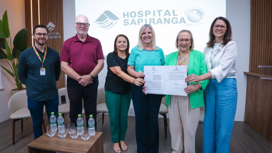 A imagem é uma fotografia de grupo que documenta a assinatura de um convênio para a compra de um mamógrafo pelo Hospital Sapiranga, no Rio Grande do Sul.

Elementos da Cena:
Grupo de Pessoas: Seis pessoas estão alinhadas no centro da imagem, sorrindo e olhando para a câmera.

No centro, duas mulheres estão segurando um documento (o convênio assinado). A mulher da esquerda veste uma blusa turquesa, e a mulher da direita veste um blazer verde-claro.

As outras quatro pessoas (dois homens nas pontas esquerda e centro-esquerda, e duas mulheres nas pontas direita e centro-direita) estão posicionadas ao lado delas, acompanhando o momento.

A maioria das pessoas usa crachás no pescoço.

Fundo (Parede/Painel):

A parede de fundo é clara, com um grande painel digital exibindo o logotipo e nome do "HOSPITAL SAPIRANGA".

À direita do logotipo do hospital, há um selo ou pequeno logotipo da "Secretaria da Saúde - OMS" ou similar.

No canto esquerdo, há um painel de madeira vertical com detalhes de textura, e o logotipo do hospital é repetido em uma placa menor ao lado.

Mobiliário (Primeiro Plano):

No canto inferior esquerdo, há uma pequena mesa de centro de madeira escura com seis garrafas de água mineral dispostas em duas filas.

Cadeiras claras e modernas são visíveis na lateral esquerda.

Conclusão Visual:
A fotografia celebra um evento institucional importante para o Hospital Sapiranga, possivelmente o anúncio ou a formalização de recursos para a aquisição de um mamógrafo, com a presença de autoridades e representantes.