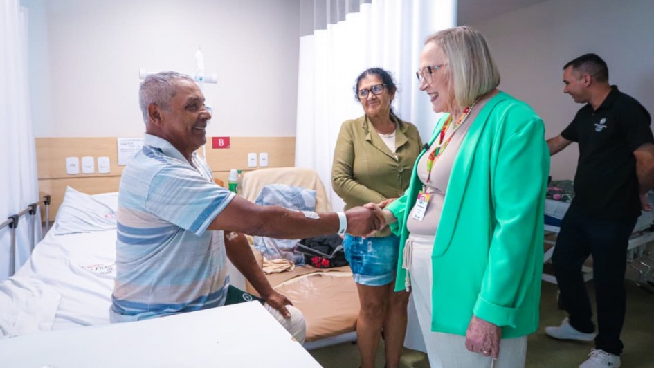 A imagem mostra um quarto hospitalar com um homem idoso sentado à beira de uma cama, estendendo a mão para cumprimentar uma mulher em pé. A cama está posicionada à esquerda, com lençóis brancos e um painel de cabeceira contendo interruptores e tomadas. Atrás do homem há uma mulher em pé, observando a cena. À direita, a secretária Arita cumprimenta o paciente. Ela veste blazer verde claro e crachá. Ao fundo, há outro homem em pé ao lado de uma segunda cama. Cortinas brancas dividem os espaços do quarto.