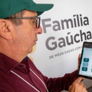 A imagem é um close-up de um homem adulto segurando e interagindo com um tablet digital, com um painel de fundo que anuncia o "Programa Família Gaúcha".

Elementos da Cena:
Homem:

Veste uma camisa polo ou social de cor vinho/bordô e um boné verde escuro.

Usa óculos de grau de armação preta e está olhando fixamente para a tela do tablet.

Sua mão direita está segurando e interagindo com a tela do dispositivo.

Tablet (Plano Central):

O tablet exibe um aplicativo ou página web com o cabeçalho "Família Gaúcha" e um menu de opções em ícones e caixas, sugerindo uma plataforma de cadastro ou acompanhamento de programas sociais.

Fundo (Painel):

A imagem tem como fundo um grande painel branco com o logotipo e nome do programa, que é "Família Gaúcha" em letras grandes, em tons de cinza.

Abaixo do nome, está o slogan "DE MÃOS DADAS COM O" (o restante do slogan é cortado).

O painel também tem símbolos gráficos coloridos e estilizados (vermelho, verde, laranja) no canto superior direito.

Conclusão Visual:
A foto ilustra uma pessoa utilizando a plataforma digital do Programa Família Gaúcha, provavelmente no contexto do processo de seleção ou acompanhamento das famílias beneficiadas, conforme o título do arquivo ("Programa Família Gaúcha finaliza seleção de 10.048 famílias").