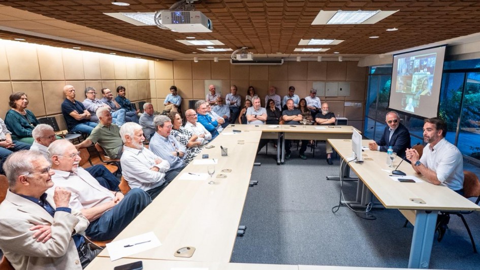 A imagem é uma fotografia em plano aberto de uma reunião ou palestra sendo realizada em uma sala de conferências. O título do arquivo sugere que é uma apresentação sobre as transformações estruturais no Rio Grande do Sul feita a economistas no Rio de Janeiro.

Elementos da Cena:
Auditório/Plateia (Plano Central e Esquerdo):

A sala está cheia, com cerca de 30 a 35 participantes sentados em cadeiras ao longo de mesas retangulares dispostas em "U" (ou em duas grandes fileiras).

Os participantes são em sua maioria homens de meia-idade ou mais velhos, vestidos com trajes casuais de negócios (camisas sociais, blazers, suéteres). Muitos estão sentados com os braços cruzados ou apoiados nas mesas, prestando atenção.

Mesa Principal (Plano Direito):

Dois palestrantes estão sentados em uma mesa na extremidade direita.

O homem à direita (vestido com camisa social branca) está falando e gesticulando com a mão direita, olhando para a plateia.

O homem à esquerda (vestido com um blazer escuro e camisa clara) está sentado de frente para a plateia, olhando para o falante.

Infraestrutura:

Teto: O teto é coberto por painéis acústicos marrons com textura, e possui diversas luminárias embutidas e um projetor multimídia.

Telão: Na parede ao fundo, à direita, há um telão de projeção exibindo slides (a imagem no telão é escura e não é claramente legível).

Paredes: As paredes laterais são cobertas por painéis acolchoados claros (bege ou marfim) para acústica.

Mesas: As mesas são de cor clara (madeira bege/marfim) e estão dispostas sobre um carpete cinza-escuro.

Conclusão Visual:
A foto captura um momento de apresentação formal e de alto nível para uma audiência especializada (economistas), provavelmente sobre políticas governamentais ou economia, com uma atmosfera de atenção concentrada.