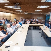 A imagem é uma fotografia em plano aberto de uma reunião ou palestra sendo realizada em uma sala de conferências. O título do arquivo sugere que é uma apresentação sobre as transformações estruturais no Rio Grande do Sul feita a economistas no Rio de Janeiro.

Elementos da Cena:
Auditório/Plateia (Plano Central e Esquerdo):

A sala está cheia, com cerca de 30 a 35 participantes sentados em cadeiras ao longo de mesas retangulares dispostas em "U" (ou em duas grandes fileiras).

Os participantes são em sua maioria homens de meia-idade ou mais velhos, vestidos com trajes casuais de negócios (camisas sociais, blazers, suéteres). Muitos estão sentados com os braços cruzados ou apoiados nas mesas, prestando atenção.

Mesa Principal (Plano Direito):

Dois palestrantes estão sentados em uma mesa na extremidade direita.

O homem à direita (vestido com camisa social branca) está falando e gesticulando com a mão direita, olhando para a plateia.

O homem à esquerda (vestido com um blazer escuro e camisa clara) está sentado de frente para a plateia, olhando para o falante.

Infraestrutura:

Teto: O teto é coberto por painéis acústicos marrons com textura, e possui diversas luminárias embutidas e um projetor multimídia.

Telão: Na parede ao fundo, à direita, há um telão de projeção exibindo slides (a imagem no telão é escura e não é claramente legível).

Paredes: As paredes laterais são cobertas por painéis acolchoados claros (bege ou marfim) para acústica.

Mesas: As mesas são de cor clara (madeira bege/marfim) e estão dispostas sobre um carpete cinza-escuro.

Conclusão Visual:
A foto captura um momento de apresentação formal e de alto nível para uma audiência especializada (economistas), provavelmente sobre políticas governamentais ou economia, com uma atmosfera de atenção concentrada.