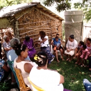 A foto mostra um grupo de pessoas reunidas em círculo ao ar livre, sentadas em cadeiras e bancos sobre a grama, à sombra de árvores. São principalmente mulheres, além de algumas crianças e duas pessoas idosas. No centro do círculo, uma mulher interage com uma senhora sentada ao seu lado. Ao fundo, há uma casa de barro com estrutura de madeira exposta e um pequeno anexo de concreto. A vegetação ao redor é densa, com árvores projetando sombra sobre o grupo.