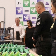 A imagem é uma fotografia em ambiente fechado (industrial) que mostra uma nova frente de trabalho para apenados na Penitenciária de Porto Alegre, em parceria com uma indústria gaúcha (logotipo Milaza ao fundo).

Elementos da Cena:
Primeiro Plano (Trabalho/Produtos):

Uma longa fileira de peças plásticas verdes (possivelmente cabos de vassouras ou outros utensílios, com formato triangular na ponta) está disposta na diagonal, pronta para serem processadas ou embaladas.

Plano Central (Ação e Pessoas):

Operário Apenado (Esquerda): Um homem (presumivelmente um apenado) está sentado atrás de uma máquina industrial (prensa ou montadora). Ele está focado na operação da máquina, processando pequenos pedaços pretos (possivelmente as peças que serão acopladas aos cabos verdes).

Autoridades/Visitantes (Centro e Direita):

Homem de Camisa Branca (Centro-Esquerda): Um homem de meia-idade, vestindo camisa social branca, está observando o trabalho na máquina.

Mulher de Preto (Centro): Uma mulher, vestindo roupa preta sem mangas, observa o processo de trabalho ao lado.

Policiais Penais (Direita): Um homem e uma mulher, ambos vestindo uniformes pretos (o homem com insígnia no ombro e a mulher com o texto "POLÍCIA PENAL" na camiseta), estão de costas para a câmera ou de perfil, supervisionando a área de trabalho.

Fundo (Institucional):

A parede de fundo é branca e exibe um painel com o logotipo da empresa parceira, "MILAZA". O logotipo é composto por corações sobrepostos em tons de verde e roxo.

Conclusão Visual:
A foto documenta a inauguração ou visita a uma oficina de trabalho dentro da Penitenciária de Porto Alegre, evidenciando a parceria entre o governo (Polícia Penal) e uma empresa privada (Milaza) na ressocialização dos apenados através do trabalho industrial.