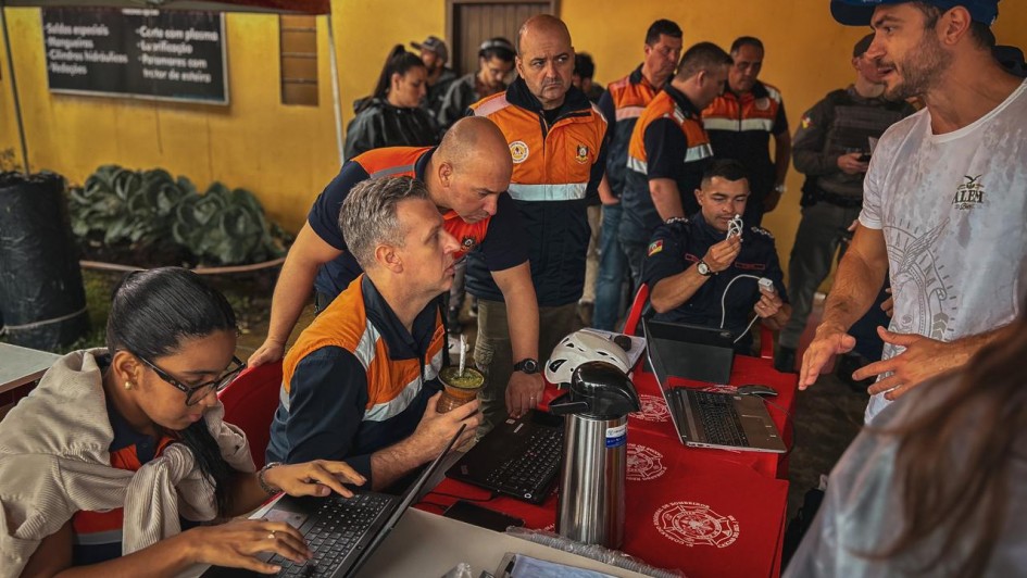 Foto de cinco homens com roupas da Defesa Civil atendendo a população numa mesa improvisada. Um homem de roupa branca gesticula enquanto fala, e os representantes do governo escutam.