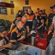 Foto de cinco homens com roupas da Defesa Civil atendendo a população numa mesa improvisada. Um homem de roupa branca gesticula enquanto fala, e os representantes do governo escutam.