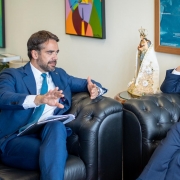 Foto de Leite e Randolfe sentados num sofá em uma sala, ambos voltados um para o outro, conversando. Leite fala e gesticula com a mão direita.