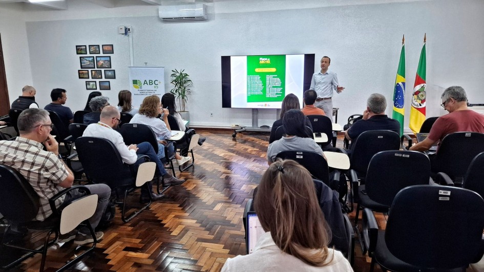 A foto mostra uma sala de reuni&otilde;es ampla, com fileiras de cadeiras pretas onde v&aacute;rias pessoas est&atilde;o sentadas voltadas para a frente. Um homem em p&eacute;, &agrave; direita da imagem, faz uma apresenta&ccedil;&atilde;o diante de uma tela grande que exibe um slide com fundo verde e textos. Ao lado da tela, est&atilde;o posicionadas duas bandeiras: a do Brasil e a do Rio Grande do Sul. Na lateral esquerda h&aacute; um banner com o texto &ldquo;ABC+ RS Plano Estadual de Agricultura de Baixa Emiss&atilde;o de Carbono&rdquo;. O piso &eacute; de madeira em padr&atilde;o escuro, e na parede ao fundo h&aacute; quadros com fotografias variadas. A sala tem ilumina&ccedil;&atilde;o clara e ar-condicionado instalado acima da tela.