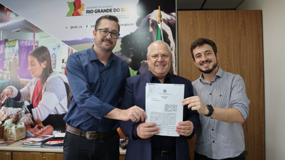 A imagem é uma fotografia de um grupo de três homens em um ambiente de escritório, em um momento de apresentação ou posse de documentos. O título do arquivo sugere que o contexto é a Consulta Popular e Emendas Parlamentares na Secretaria de Desenvolvimento Rural (SDR).

Elementos da Cena:
Pessoas: Três homens adultos estão posicionados lado a lado, voltados para a câmera, sorrindo e segurando um documento.

Homem do Centro: É o mais velho, usa um blazer azul-marinho sobre uma camisa social clara. Ele está segurando o documento oficial (provavelmente um convênio ou termo de acordo) com as duas mãos. Ele é ladeado pelos outros dois.

Homem da Esquerda: Veste uma camisa social azul escura e óculos de grau. Ele toca o documento com a mão direita, junto ao homem central.

Homem da Direita: Veste uma camisa social cinza de listras verticais e um relógio preto no pulso esquerdo. Ele toca o documento com a mão esquerda.

Fundo Institucional:

A parede de fundo é predominantemente branca e bege.

Há um grande painel de comunicação na parede, com o logotipo do Governo do Estado do Rio Grande do Sul, o texto "SECRETARIA DE DESENVOLVIMENTO RURAL" (SDR) e o endereço web sdr.rs.gov.br.

O painel exibe imagens fotográficas de fundo que representam a agricultura e o desenvolvimento rural (como uma mulher trabalhando e uma imagem parcial de uma vaca).

O lado direito do fundo é uma parede de madeira clara com textura de painéis verticais.

Conclusão Visual:
A foto documenta a formalização de um termo (relacionado à Consulta Popular ou Emendas Parlamentares) na Secretaria de Desenvolvimento Rural do Rio Grande do Sul, com a participação de três representantes.