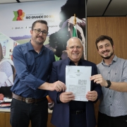 A imagem é uma fotografia de um grupo de três homens em um ambiente de escritório, em um momento de apresentação ou posse de documentos. O título do arquivo sugere que o contexto é a Consulta Popular e Emendas Parlamentares na Secretaria de Desenvolvimento Rural (SDR).

Elementos da Cena:
Pessoas: Três homens adultos estão posicionados lado a lado, voltados para a câmera, sorrindo e segurando um documento.

Homem do Centro: É o mais velho, usa um blazer azul-marinho sobre uma camisa social clara. Ele está segurando o documento oficial (provavelmente um convênio ou termo de acordo) com as duas mãos. Ele é ladeado pelos outros dois.

Homem da Esquerda: Veste uma camisa social azul escura e óculos de grau. Ele toca o documento com a mão direita, junto ao homem central.

Homem da Direita: Veste uma camisa social cinza de listras verticais e um relógio preto no pulso esquerdo. Ele toca o documento com a mão esquerda.

Fundo Institucional:

A parede de fundo é predominantemente branca e bege.

Há um grande painel de comunicação na parede, com o logotipo do Governo do Estado do Rio Grande do Sul, o texto "SECRETARIA DE DESENVOLVIMENTO RURAL" (SDR) e o endereço web sdr.rs.gov.br.

O painel exibe imagens fotográficas de fundo que representam a agricultura e o desenvolvimento rural (como uma mulher trabalhando e uma imagem parcial de uma vaca).

O lado direito do fundo é uma parede de madeira clara com textura de painéis verticais.

Conclusão Visual:
A foto documenta a formalização de um termo (relacionado à Consulta Popular ou Emendas Parlamentares) na Secretaria de Desenvolvimento Rural do Rio Grande do Sul, com a participação de três representantes.