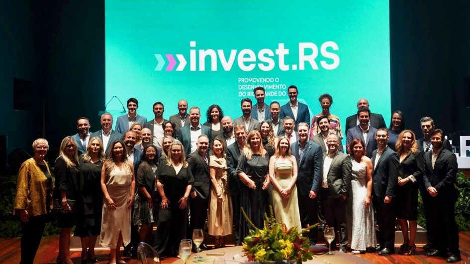 A foto mostra um grande grupo de pessoas posando para uma foto oficial em frente a um tel&atilde;o azul-esverdeado com o logotipo &ldquo;invest.RS&rdquo; e a frase &ldquo;Promovendo o desenvolvimento do Rio Grande do Sul&rdquo;. O grupo &eacute; composto por homens e mulheres organizados em v&aacute;rias fileiras. Em primeiro plano, h&aacute; mesas com arranjos florais e ta&ccedil;as com bebida. O ambiente &eacute; um audit&oacute;rio iluminado com luz c&ecirc;nica.