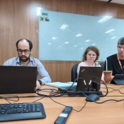 Foto de três pessoas sentadas junto a uma mesa, em frente a notebooks.