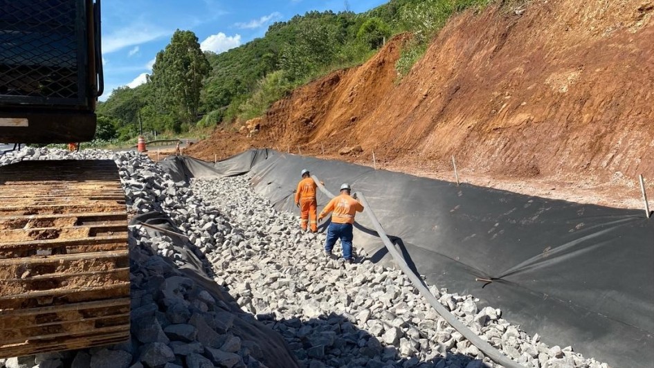 A imagem é uma fotografia em plano fechado que captura uma frente de trabalho em uma rodovia (VRS-826, entre Feliz e Alto Feliz), focada na contenção de encostas e recuperação da estrada.

Elementos da Cena:
Primeiro Plano (Trabalho):

O lado esquerdo é dominado pela parte inferior de uma máquina pesada (provavelmente uma escavadeira ou trator), com sua esteira (rastros) de cor marrom-avermelhada e ferrugem visível.

O solo na área de trabalho está coberto por uma grande quantidade de pedras soltas (rachão) de cor cinza-clara, que formam o leito da recuperação.

Plano Central (Obras):

Uma manta geotêxtil preta está estendida sobre o terreno, cobrindo o talude (a encosta) e parte da área onde as pedras estão sendo colocadas. Essa manta serve para contenção e drenagem.

Dois operários estão presentes:

Um está de costas (à esquerda), vestindo um macacão de trabalho laranja e um capacete branco.

O outro (à direita) veste uma camisa laranja e calça azul-escura, e está caminhando sobre as pedras.

Um tubo de drenagem preto está posicionado sobre as pedras, próximo à manta.

Plano de Fundo (Encosta):

À direita, a encosta do morro é alta, com terra em tom vermelho-ferrugem exposta, indicando um deslizamento ou trabalho de corte recente.

A parte superior da encosta e o fundo da imagem são cobertos por vegetação densa de cor verde (mata nativa ou arbustos).

Céu: O céu está azul e claro, com luz solar intensa, indicando um dia de tempo bom para a realização das obras.

Conclusão Visual:
A foto evidencia o trabalho em andamento de recuperação da rodovia, com foco na estabilização do talude e drenagem, após danos causados por deslizamentos ou erosão (conforme sugerido pela terra vermelha exposta na encosta).