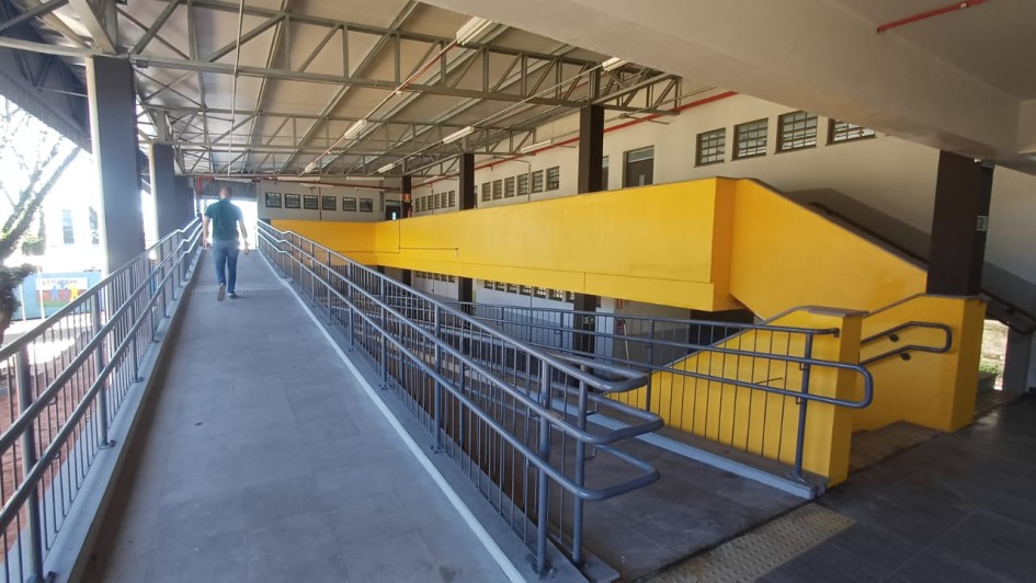 ♿ Descrição da Imagem para Acessibilidade
Esta é uma fotografia em plano médio de uma rampa de acesso para pedestres e cadeirantes, localizada em uma área coberta, possivelmente na entrada ou dentro de um prédio escolar ou público.

A rampa, no centro e à esquerda, possui piso cinza escuro e uma inclinação suave. É equipada com corrimãos duplos de metal cinza (guarda-corpos) em ambos os lados, garantindo acessibilidade e segurança. No lado direito da rampa, o corrimão é contínuo e segue a inclinação.

No centro da imagem, uma grande parede lateral, que acompanha a rampa, é pintada em um chamativo e sólido amarelo vivo. A estrutura amarela se eleva e termina em uma escada na extremidade direita.

Em primeiro plano, na base da rampa, notam-se faixas de piso tátil de alerta na cor amarela, indicando o início da inclinação.

Um pedestre (um homem vestindo camiseta azul escura e calças claras) está subindo a rampa, caminhando de costas para o observador, no terço esquerdo da imagem.

A cobertura é composta por uma estrutura metálica de treliças e telhado translúcido ou de metal claro, que permite a entrada de luz natural. O teto possui luminárias embutidas ou suspensas.

Ao fundo, à direita, a estrutura amarela se conecta a uma parede de um edifício de cor marrom (ou revestimento escuro), com diversas janelas horizontais e pequenas em sua parte superior, alinhadas em fileiras.