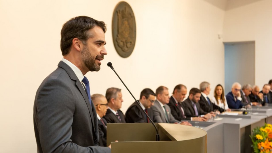Esta é uma fotografia de um evento solene ou cerimônia formal em um ambiente interno.

Em primeiro plano e à esquerda, está o Governador Eduardo Leite, em perfil (meio-corpo), discursando em um pódio.

Ele veste um terno cinza escuro, camisa social branca e gravata azul.

Ele tem barba e cabelo escuros e está olhando ligeiramente para a frente e para a direita.

Suas mãos estão visíveis, apoiadas no pódio.

O pódio é moderno, de formato curvo e cor bege ou cáqui escura. Um microfone de pedestal preto está posicionado para captar a fala do governador.

Ao fundo, no centro, uma longa mesa cinza se estende para a direita, onde estão sentadas diversas autoridades e convidados. Eles estão vestidos com trajes formais (ternos escuros e togas pretas, sugerindo um evento de posse ou tribunal).

A maioria das pessoas sentadas são homens de meia-idade ou mais velhos, mas há algumas mulheres.

Eles estão olhando na direção do orador.

A decoração da sala é minimalista:

As paredes são lisas e de cor bege ou creme clara.

No centro da parede de fundo, acima da linha das cabeças, há um grande emblema ou brasão redondo e escuro (marrom ou bronze), possivelmente o símbolo do Tribunal de Contas do Estado (TCE).

No canto inferior direito, uma composição floral com predominância de flores laranjas e amarelas está parcialmente visível.

A iluminação é quente e concentra-se no orador em primeiro plano, enquanto o fundo está em sombra suave.