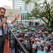 Foto de Leite falando ao microfone, em cima de um trio elétrico. Há uma grande concentração de pessoas na praça, acompanhando em capas de chuva.