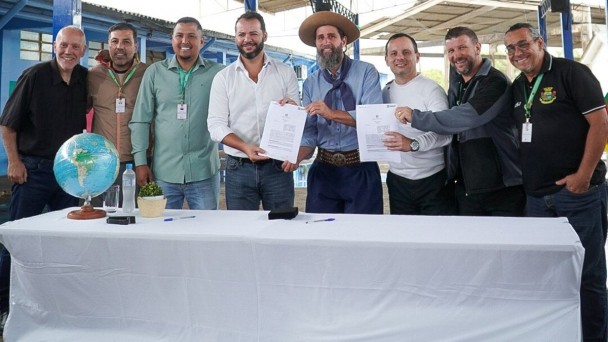 ⚽ Descri&ccedil;&atilde;o da Imagem para Acessibilidade
Esta &eacute; uma fotografia em plano m&eacute;dio de um grupo de oito homens posando em um ambiente externo (possivelmente coberto, como um gin&aacute;sio ou galp&atilde;o), celebrando a assinatura de um documento. O evento &eacute; a assinatura de um conv&ecirc;nio para revitaliza&ccedil;&atilde;o de uma quadra esportiva em Alvorada.

O Grupo (da esquerda para a direita):

Homem 1: Sorridente, veste camisa preta e cal&ccedil;as escuras.

Homem 2: Veste uma jaqueta marrom clara (c&aacute;qui) sobre uma camisa escura e cal&ccedil;as escuras.

Homem 3: Veste uma camisa polo verde-clara, cal&ccedil;as jeans e um crach&aacute; pendurado no pesco&ccedil;o. Ele tem barba e est&aacute; sorrindo.

Homem 4 (no centro): Veste camisa social branca (com as mangas arrega&ccedil;adas), cal&ccedil;as jeans escuras. Ele tem barba e est&aacute; segurando um dos documentos abertos. Ele est&aacute; sorrindo.

Homem 5 (em destaque): No centro, veste trajes t&iacute;picos ga&uacute;chos (pilcha): bombacha azul-escura, len&ccedil;o azul no pesco&ccedil;o, camisa clara e um chap&eacute;u de abas largas (estilo ga&uacute;cho). Ele tem barba longa e est&aacute; sorrindo, segurando o outro documento aberto.

Homem 6: Veste uma camisa branca de manga longa e cal&ccedil;as escuras. Ele est&aacute; segurando uma caneta ou apontando para um dos documentos.

Homem 7: Veste camisa polo cinza escura, cal&ccedil;as escuras. Ele est&aacute; com a m&atilde;o no ombro do Homem 6, sorrindo.

Homem 8 (extrema direita): Veste uma camisa polo preta com bordados ou logotipo de um munic&iacute;pio ou organiza&ccedil;&atilde;o (possivelmente Alvorada) e &oacute;culos. Ele est&aacute; sorrindo.

O Fundo e os Objetos na Mesa:

Mesa: O grupo est&aacute; atr&aacute;s de uma mesa longa coberta por uma toalha branca, onde os documentos foram assinados.

Objetos: Sobre a mesa, no lado esquerdo, h&aacute; um globo terrestre em um pedestal, garrafas de &aacute;gua e canetas pretas e azuis.

Fundo: O fundo &eacute; um ambiente aberto, com estruturas met&aacute;licas e paredes de madeira ou metal pintadas de azul claro e branco, indicando um galp&atilde;o, gin&aacute;sio ou &aacute;rea de lazer coberta.

A imagem &eacute; luminosa e capta a formaliza&ccedil;&atilde;o de um acordo com a presen&ccedil;a de representantes municipais, incluindo uma figura que representa a cultura local.