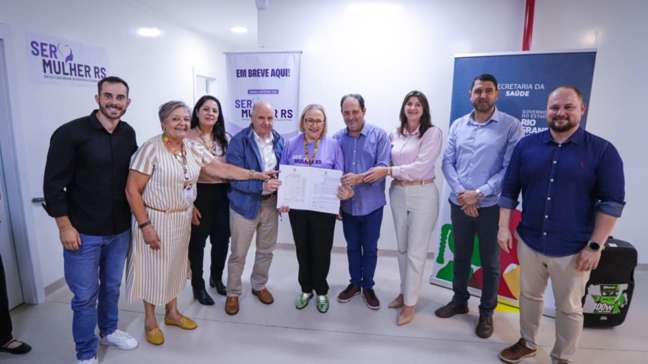 
Uma fotografia colorida captura um grupo de nove pessoas adultas, em pé, posando juntas em um ambiente interno bem iluminado, provavelmente uma instalação de saúde ou evento governamental.

Pessoas e Ação Central:
No centro da imagem, quatro pessoas estão segurando um ou mais documentos juntos, em um ato que simboliza uma assinatura, acordo ou entrega oficial.

Uma mulher de cabelos loiros curtos, vestindo uma blusa roxa, está no centro exato, segurando os papéis com ambas as mãos.

Um homem careca com camisa azul clara está à esquerda dela, também tocando os documentos.

Um homem com camisa azul escura está à direita dela, também tocando os documentos.

À extrema esquerda do grupo central, uma mulher de cabelos grisalhos, vestindo um vestido listrado bege e amarelo, sorri amplamente e aponta para os documentos ou para o grupo.

Outros Membros do Grupo:
À esquerda, um jovem de camisa preta e calças jeans está sorrindo.

Entre o jovem e a mulher de vestido listrado, estão uma mulher e um homem, ambos sorrindo.

À direita do grupo central, uma mulher de camisa rosa claro e calças brancas, e dois homens, um de camisa azul de botões e outro de camisa polo marrom, completam a formação, todos sorrindo e olhando para frente.

Fundo e Contexto:
O ambiente é de paredes claras, e há banners de propaganda ou informativos no fundo que fornecem contexto:

À esquerda, um banner roxo tem o texto "SER MULHER RS" com um símbolo.

Acima do grupo central, um cartaz azul diz "EM BREVE AQUI!" e, abaixo, o logo do SER MULHER RS se repete.

À direita, um banner azul escuro com o logo do GOVERNO DO ESTADO DO RIO GRANDE DO SUL e o texto "SECRETARIA DA SAÚDE" (indicando o local ou a natureza do evento).

O evento parece ser a celebração de uma parceria ou habilitação de um serviço de saúde.