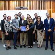 Um grupo de dez pessoas posa para foto com um documento em mãos em um ambient de uma sala formal.