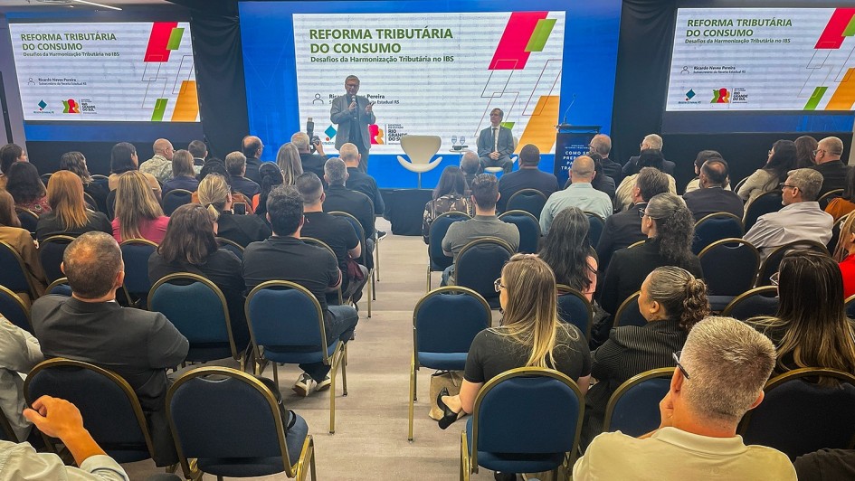 Foto mostra sala cheia, de pessoas sentadas de costas para a câmera. Elas estão olhando o palestrante que está em pé com microfone na mão. Ele veste um terno cinza claro. Ao fundo, três telões com slide sobre reforma tributária.