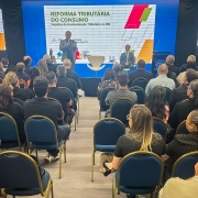 Foto mostra sala cheia, de pessoas sentadas de costas para a câmera. Elas estão olhando o palestrante que está em pé com microfone na mão. Ele veste um terno cinza claro. Ao fundo, três telões com slide sobre reforma tributária.