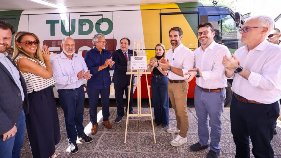 Foto externa mostrando um grupo de pessoas em pé diante de um veículo grande adesivado com as cores verde, amarela e vermelha e os dizeres “TUDO FÁCIL”. No centro, há um cavalete com placa inaugurada. Todos aplaudem, entre eles o governador Eduardo Leite.