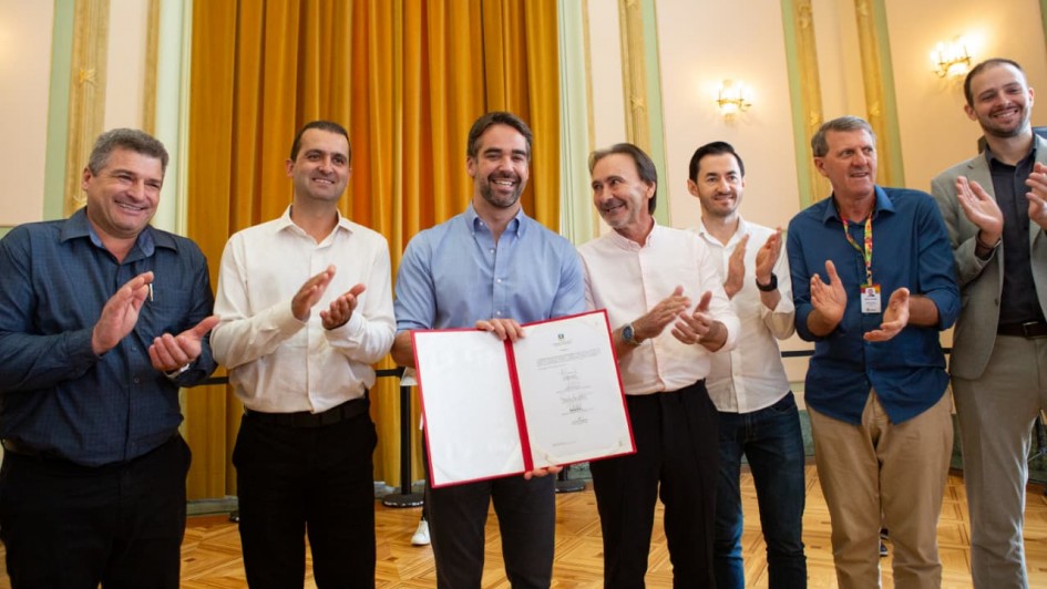 Foto em ambiente interno de um salão amplo e clássico, com cortinas longas em tom dourado ao fundo e paredes claras com detalhes decorativos. No centro da imagem, o governador Eduardo Leite segura um documento aberto, exibindo seu conteúdo para a câmera. Ele veste camisa social de manga longa em tom claro e sorri. Ao seu redor, há outros homens adultos em pé, dispostos lado a lado, aplaudindo. Todos vestem roupas sociais ou semiformais. O piso é de madeira e a iluminação é quente, proveniente de lustres e arandelas nas paredes.