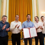 Foto em ambiente interno de um salão amplo e clássico, com cortinas longas em tom dourado ao fundo e paredes claras com detalhes decorativos. No centro da imagem, o governador Eduardo Leite segura um documento aberto, exibindo seu conteúdo para a câmera. Ele veste camisa social de manga longa em tom claro e sorri. Ao seu redor, há outros homens adultos em pé, dispostos lado a lado, aplaudindo. Todos vestem roupas sociais ou semiformais. O piso é de madeira e a iluminação é quente, proveniente de lustres e arandelas nas paredes.