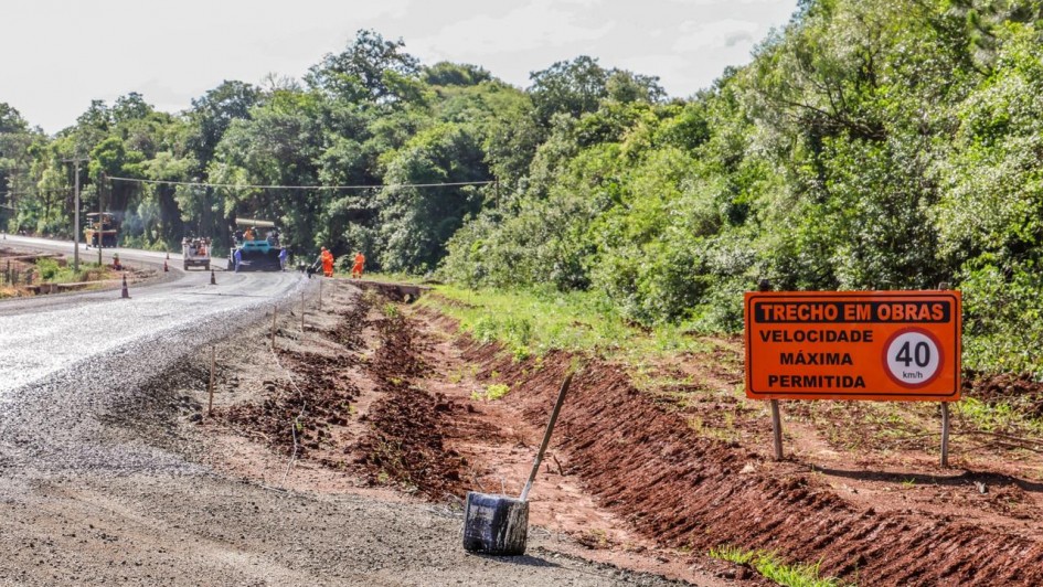 
Esta é uma fotografia em plano geral de um trecho de estrada em obras, sob luz solar.

O primeiro plano e o centro da imagem mostram a estrada em construção ou recapeamento.

O lado esquerdo é composto por cascalho solto e irregular, indicando o leito da via em fase inicial ou intermediária da obra.

À direita da estrada, há uma faixa de terra marrom-avermelhada (saibro), escavada para a drenagem ou alargamento da via.

Um balde preto (possivelmente para ferramentas ou entulho) está tombado no meio desta faixa de terra, com o cabo de uma pá ou ferramenta apoiado na terra ao lado.

Em segundo plano, à esquerda, a via principal pavimentada (asfalto ou concreto) é visível, com veículos e equipamentos de construção:

Dois veículos de cor laranja e azul, que parecem ser máquinas de pavimentação (rolo compressor e/ou vibroacabadora), estão estacionados ou em operação.

Alguns trabalhadores, vestindo coletes de segurança laranja, estão próximos às máquinas.

No lado direito, em destaque, um sinal de trânsito temporário (placa de obras) está fixado na lateral da estrada.

A placa é retangular, de cor laranja vibrante, com texto em branco e preto.

O texto diz: "TRECHO EM OBRAS"

Abaixo, lê-se: "VELOCIDADE MÁXIMA PERMITIDA"

Um círculo branco com borda vermelha ao lado indica a velocidade de "40 km/h".

O fundo da imagem é dominado por uma vegetação densa de floresta ou mata, com árvores altas e folhagem verde escura, típica de uma área rural.

A imagem transmite o ambiente de uma obra rodoviária em andamento, como a vistoria na ERS-403, entre Rio Pardo e Cachoeira do Sul, conforme o nome do arquivo.