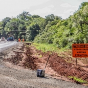 
Esta é uma fotografia em plano geral de um trecho de estrada em obras, sob luz solar.

O primeiro plano e o centro da imagem mostram a estrada em construção ou recapeamento.

O lado esquerdo é composto por cascalho solto e irregular, indicando o leito da via em fase inicial ou intermediária da obra.

À direita da estrada, há uma faixa de terra marrom-avermelhada (saibro), escavada para a drenagem ou alargamento da via.

Um balde preto (possivelmente para ferramentas ou entulho) está tombado no meio desta faixa de terra, com o cabo de uma pá ou ferramenta apoiado na terra ao lado.

Em segundo plano, à esquerda, a via principal pavimentada (asfalto ou concreto) é visível, com veículos e equipamentos de construção:

Dois veículos de cor laranja e azul, que parecem ser máquinas de pavimentação (rolo compressor e/ou vibroacabadora), estão estacionados ou em operação.

Alguns trabalhadores, vestindo coletes de segurança laranja, estão próximos às máquinas.

No lado direito, em destaque, um sinal de trânsito temporário (placa de obras) está fixado na lateral da estrada.

A placa é retangular, de cor laranja vibrante, com texto em branco e preto.

O texto diz: "TRECHO EM OBRAS"

Abaixo, lê-se: "VELOCIDADE MÁXIMA PERMITIDA"

Um círculo branco com borda vermelha ao lado indica a velocidade de "40 km/h".

O fundo da imagem é dominado por uma vegetação densa de floresta ou mata, com árvores altas e folhagem verde escura, típica de uma área rural.

A imagem transmite o ambiente de uma obra rodoviária em andamento, como a vistoria na ERS-403, entre Rio Pardo e Cachoeira do Sul, conforme o nome do arquivo.