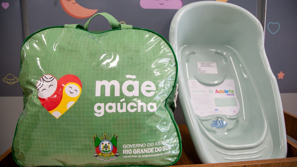 Foto mostra, sobre um m&oacute;vel de madeira, um kit de maternidade. &Agrave; esquerda, h&aacute; uma bolsa verde com al&ccedil;a, transparente na parte frontal, com o logotipo &ldquo;m&atilde;e ga&uacute;cha&rdquo; e o bras&atilde;o do Governo do Estado do Rio Grande do Sul, identificando a Secretaria de Desenvolvimento Social. Ao lado da bolsa, &agrave; direita, aparece uma banheira infantil de pl&aacute;stico na cor verde-claro, com etiqueta do fabricante vis&iacute;vel no fundo interno. O fundo da imagem &eacute; um painel decorativo com desenhos infantis, como lua, nuvem e cora&ccedil;&atilde;o.