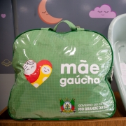 Foto mostra, sobre um m&oacute;vel de madeira, um kit de maternidade. &Agrave; esquerda, h&aacute; uma bolsa verde com al&ccedil;a, transparente na parte frontal, com o logotipo &ldquo;m&atilde;e ga&uacute;cha&rdquo; e o bras&atilde;o do Governo do Estado do Rio Grande do Sul, identificando a Secretaria de Desenvolvimento Social. Ao lado da bolsa, &agrave; direita, aparece uma banheira infantil de pl&aacute;stico na cor verde-claro, com etiqueta do fabricante vis&iacute;vel no fundo interno. O fundo da imagem &eacute; um painel decorativo com desenhos infantis, como lua, nuvem e cora&ccedil;&atilde;o.